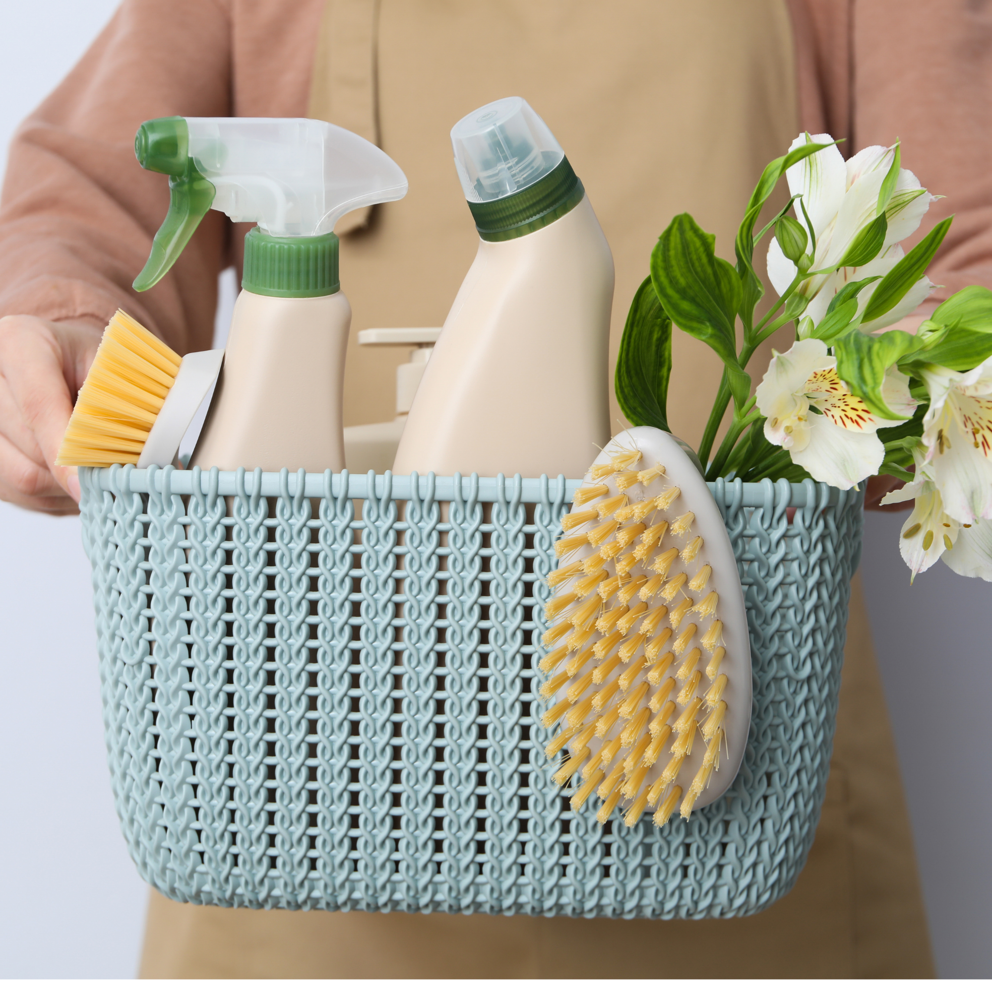 Person holding a basket with cleaning supplies and white flowers.