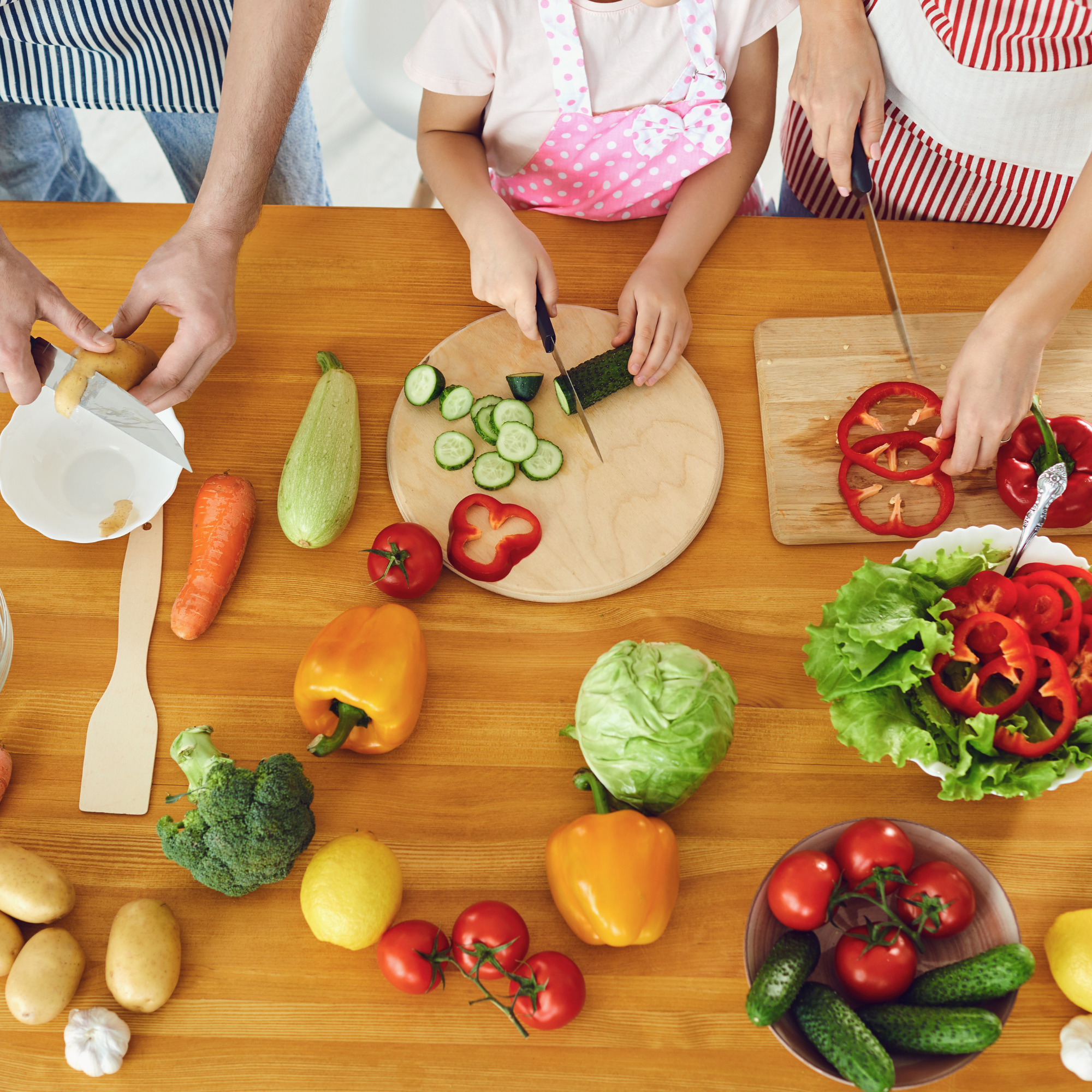 Photo of assorted vegetables on a wooden counter with 3 sets of hands chopping.